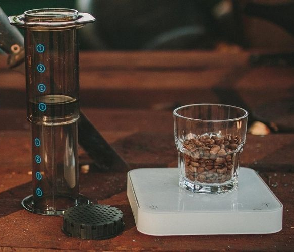 Cafeteira aeropress ao centro com um copo ao lado direito e um saco de café a esquerda