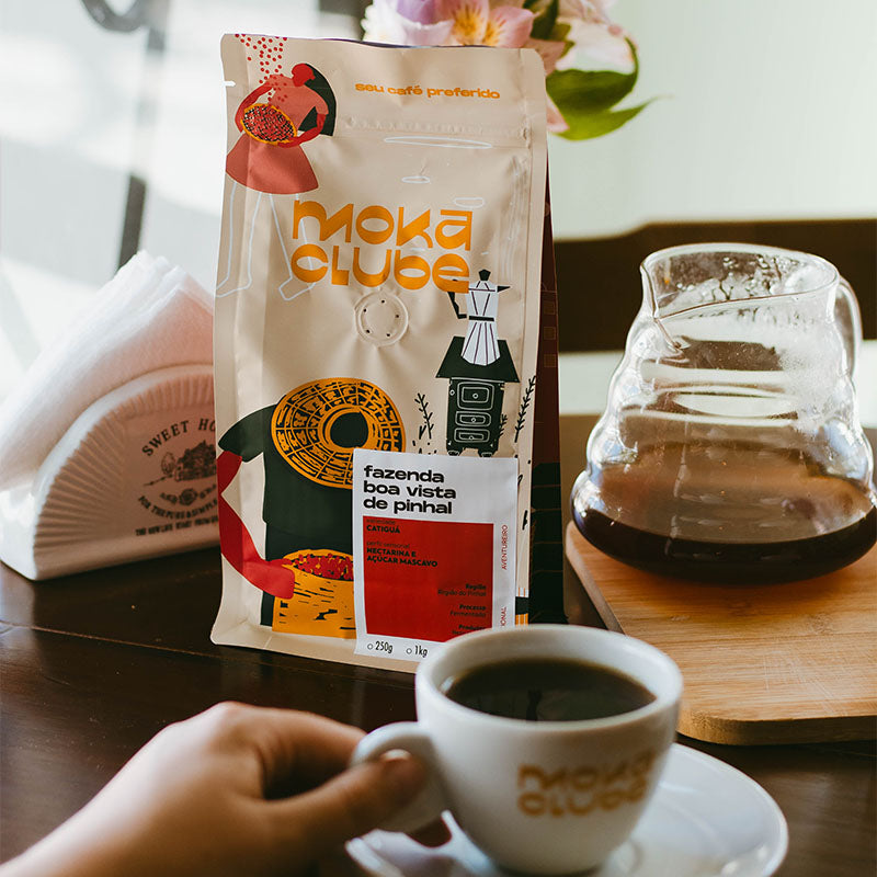 Mesa com uma xícara branca do moka clube, uma jarra de viro com café, pacote de café, flores e guardanapos
