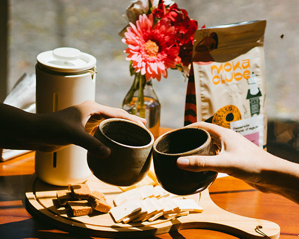 Um brinde com copos de cerâmica, tábua de frios, prensa francesa, flores e pacote de café