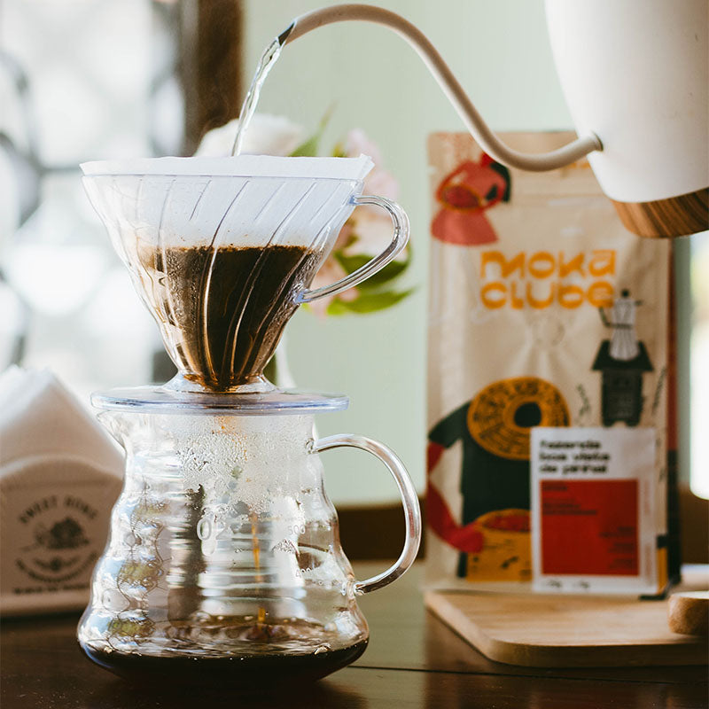Chaleira despejando água no café em um kit V60 ao fundo pacote de café, flores e guardanapos