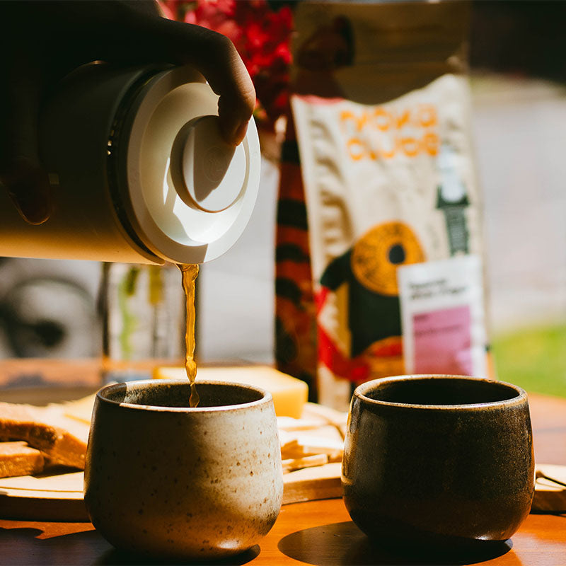 Uma prensa Francesa Timemore servindo café em copos de cirâmica ao fundo pacote de café
