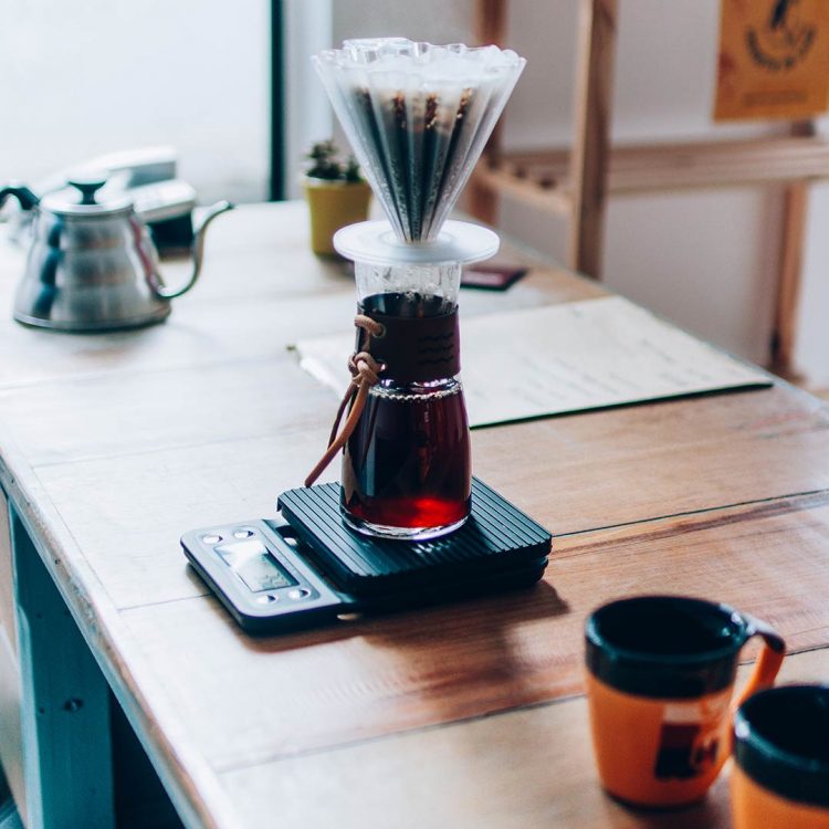 Uma mesa com uma chaleira hário ao fundo, um decanter com porta filtro em cima de uma balança e xicaras laranjas