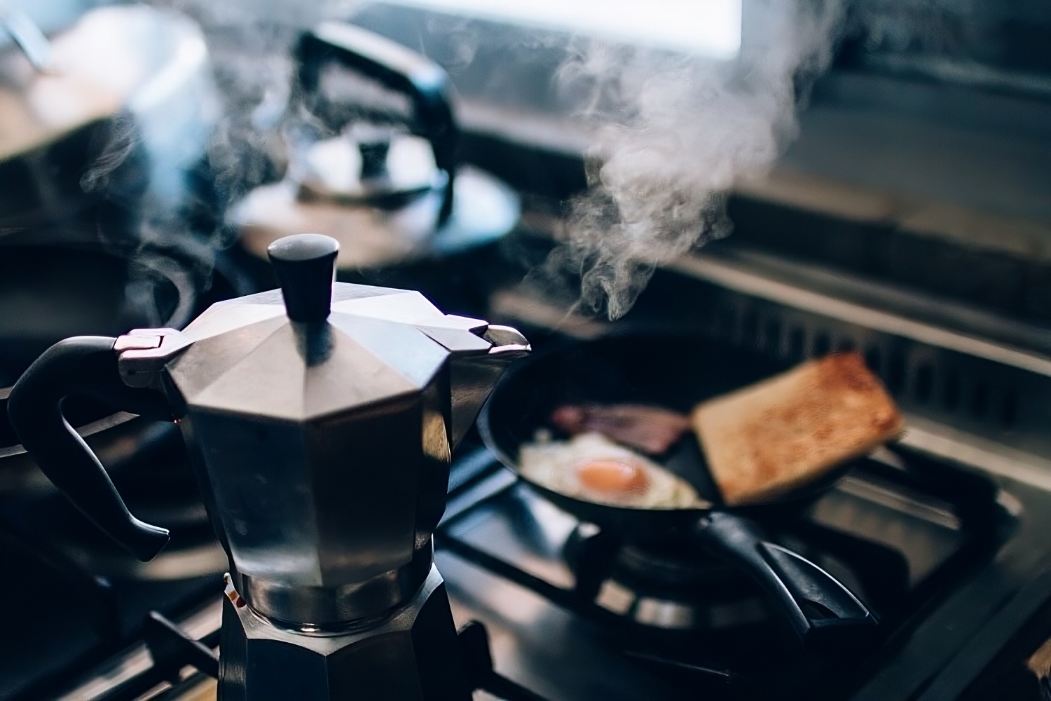 Café sendo preparado em cafeteira italiana no fogão com frigideira e chaleira no fundo