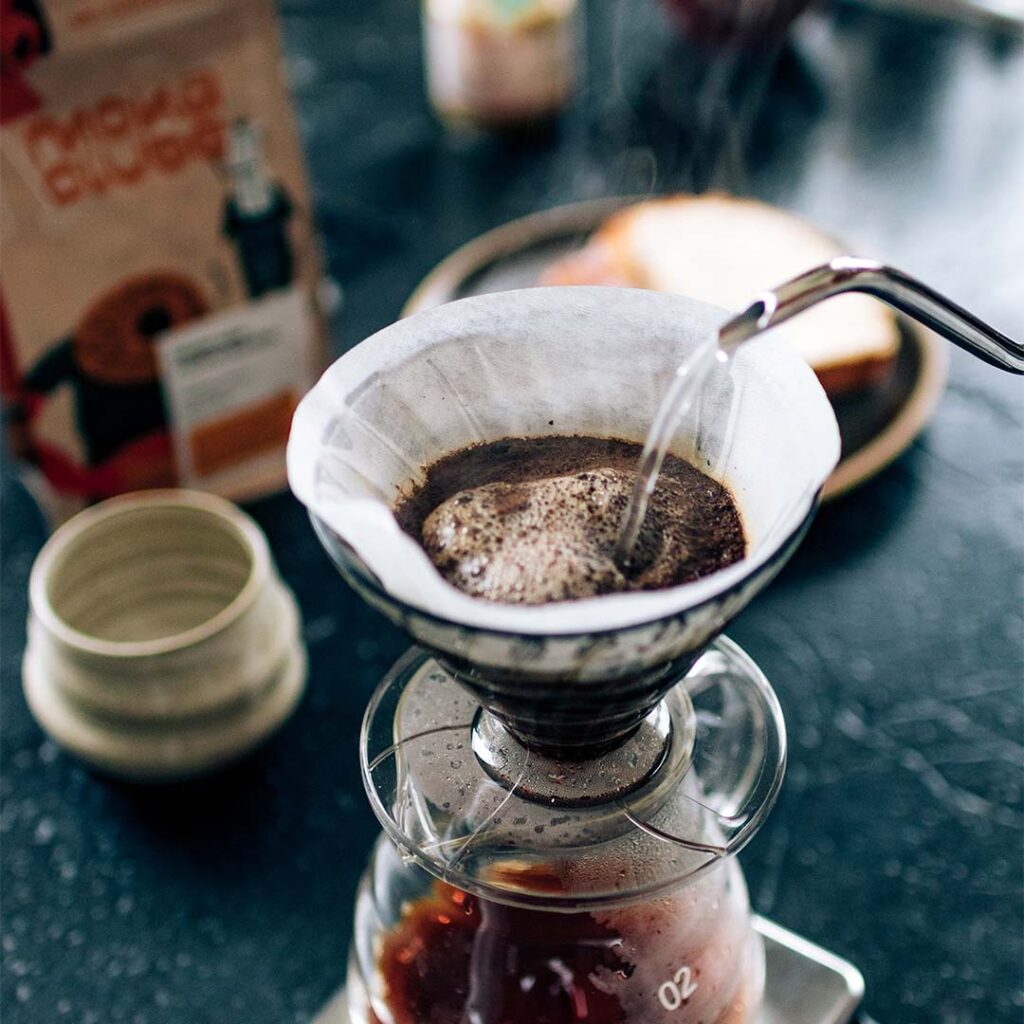 Café sendo coado em porta filtro em cima e mesa com copo de cerâmica, pacote de café e prato com pão